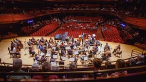 Photo from above and behind the Anderson Hall stage showing the practicing orchestra and a mostly empty house of red seats