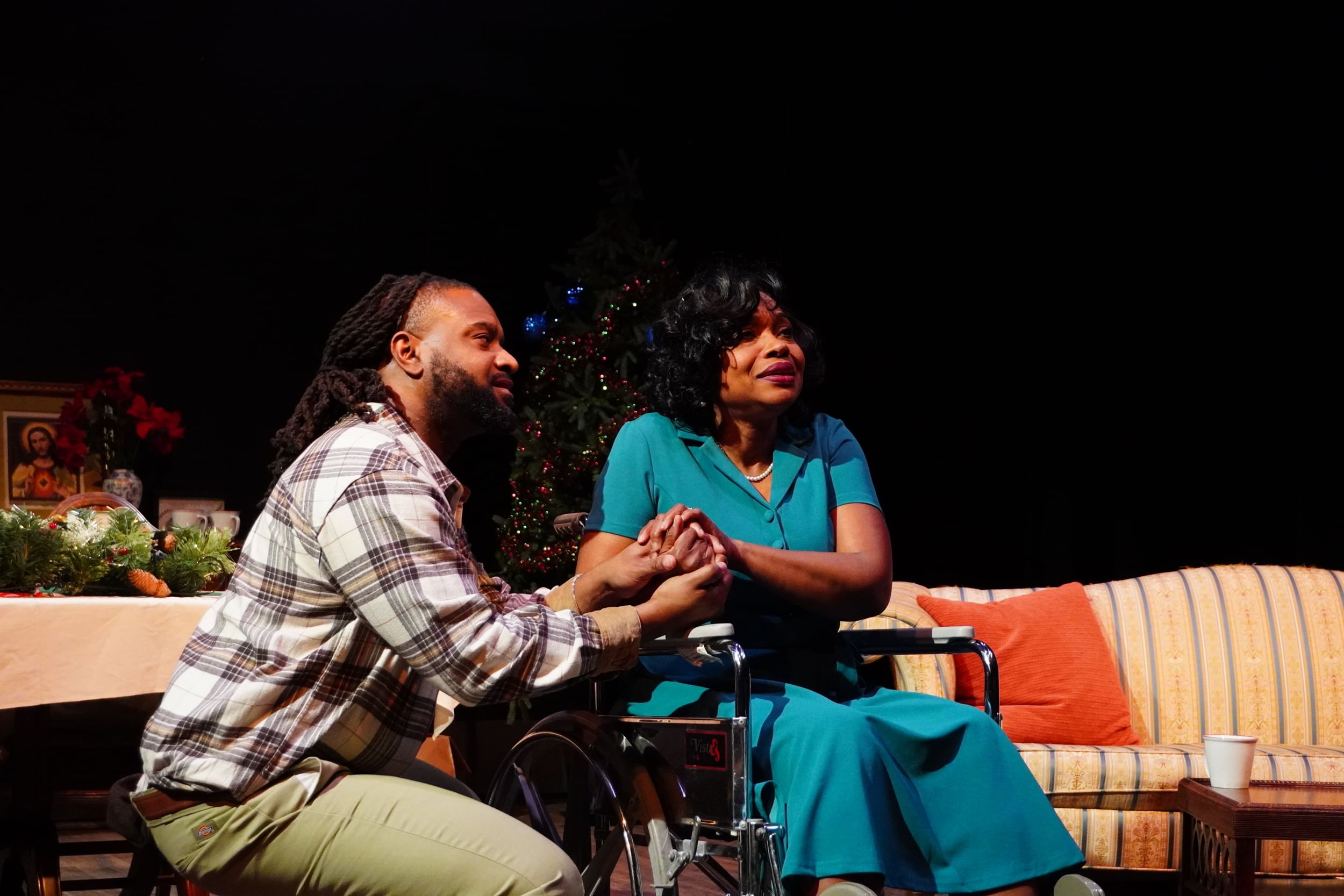 Grant, a Black man in a plaid shirt, clasps hands with Booker, a Black woman in a turquoise dress, sitting in a wheelchair.