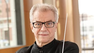 Vänskä, a 72-year-old white man with short gray hair & glasses, looks directly at you with arms folded, a baton in one hand