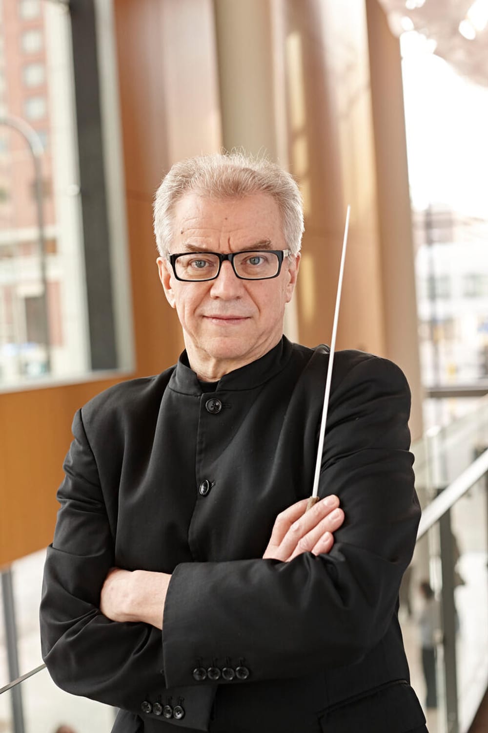 Vänskä, a 72-year-old white man with short gray hair & glasses, looks directly at you with arms folded, a baton in one hand