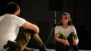 Dana, with his back to us in a white tee, boots, fatigues and gun, sits facing Gillespie, in similar clothes, looking worried