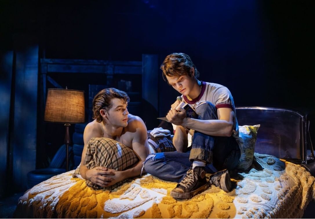 Two young white men in worn 1960s-style denim, on a bed with a worn yellow & white cover, study a book with tender attention