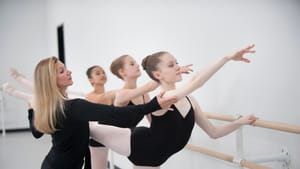 Santoro adjusts a student’s posture, three students in leotards in the rehearsal space by a ballet barre.