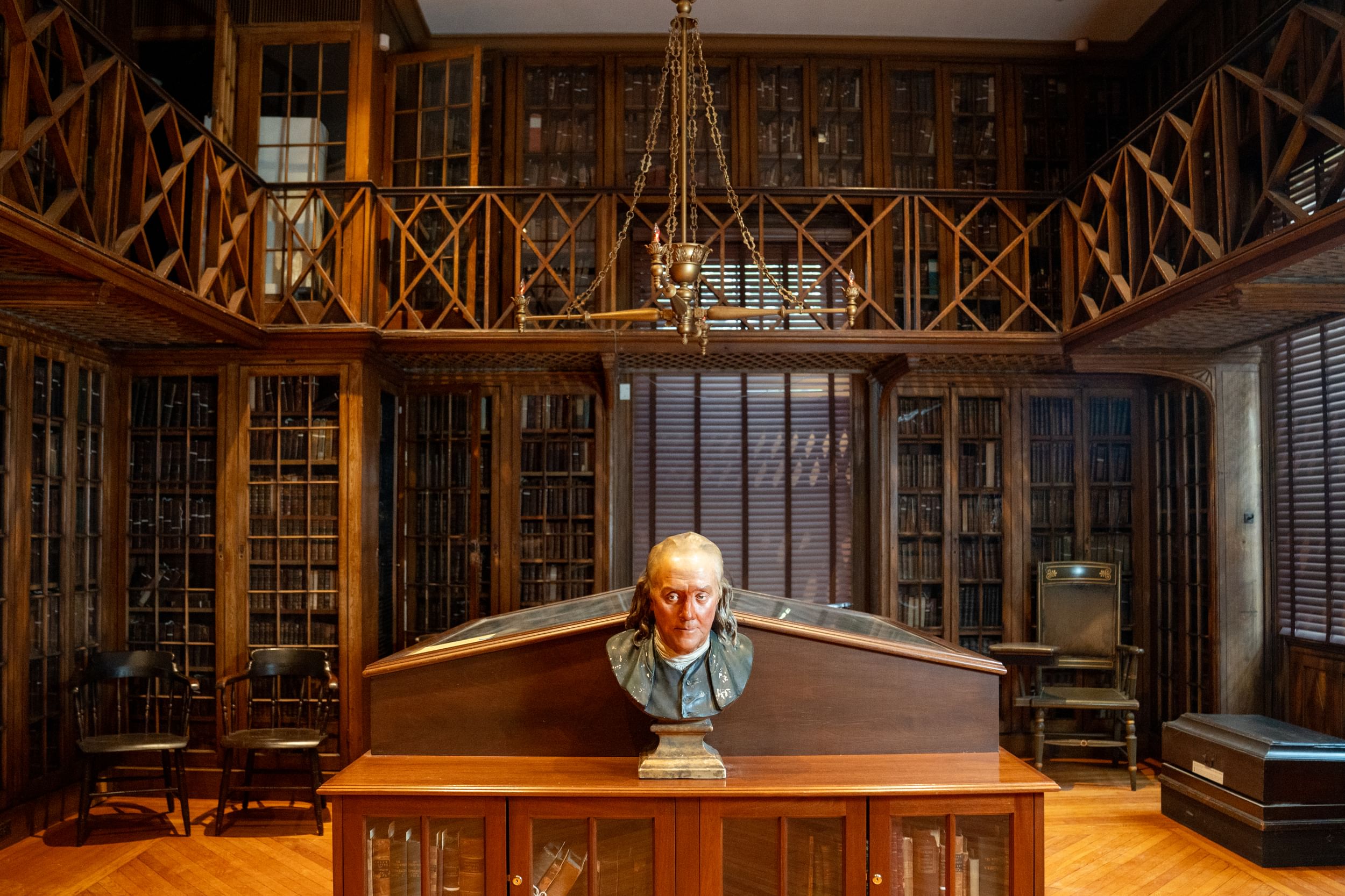 An ornate two-tiered, wood-paneled 18th-century library with glass cases. A painted bust of Ben Franklin sits at center.