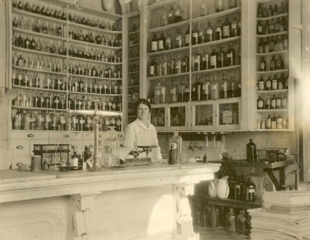 Vintage sepia photo of a woman in white uniform in front of tall shelves of remedies in little glass jars.