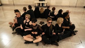 15 people, all in black, sit in a circle on the floor facing outward, arms linked, a pile of cell phones in the middle.