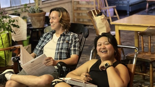 Robinson, in a plaid shirt, and Alvarez, in a black tank, sit smiling in chairs holding scripts at a living-room rehearsal
