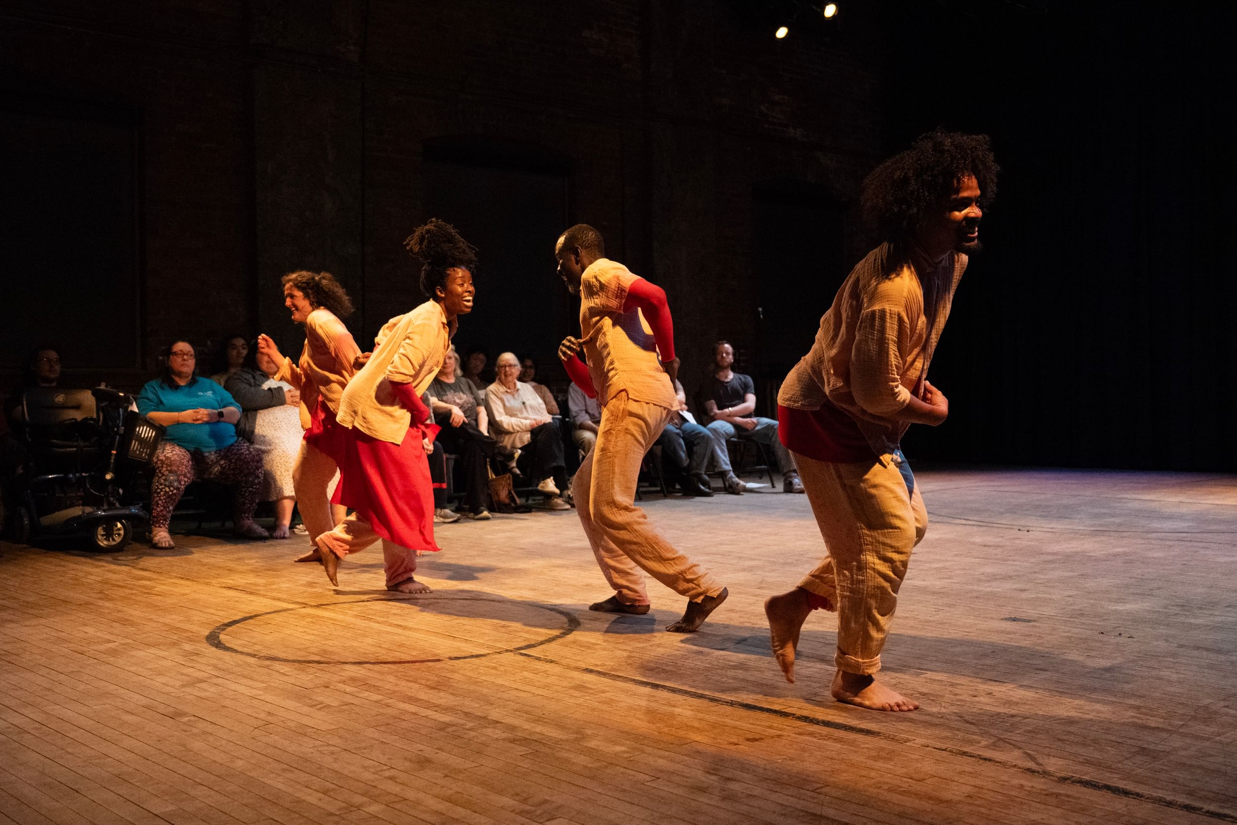 Four dancers in loose, pale clothes dance joyfully in a line on an old gym floor as the audience watches behind them.