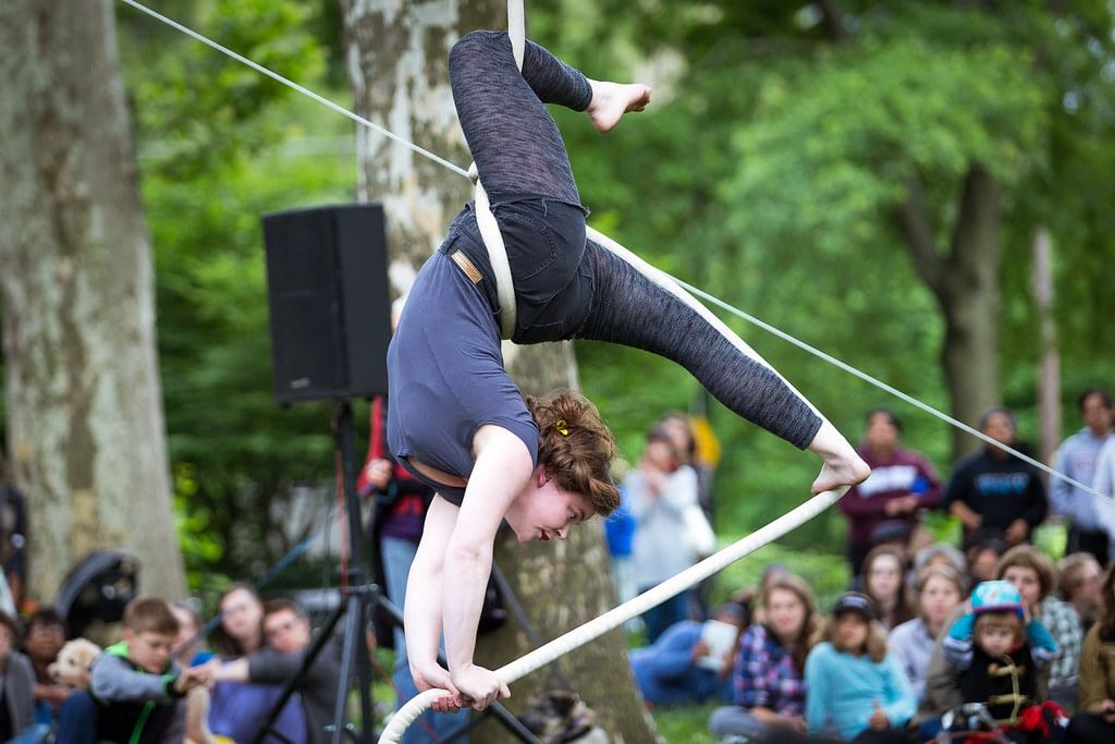 A woman doing an aerial performance in a summer park strikes a complex triangular pose with a rope while the audience watches