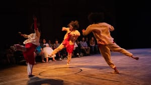 Three PDP dancers, each on one leg in the middle of a dynamic tilting move, perform in a venue with an old gym floor.