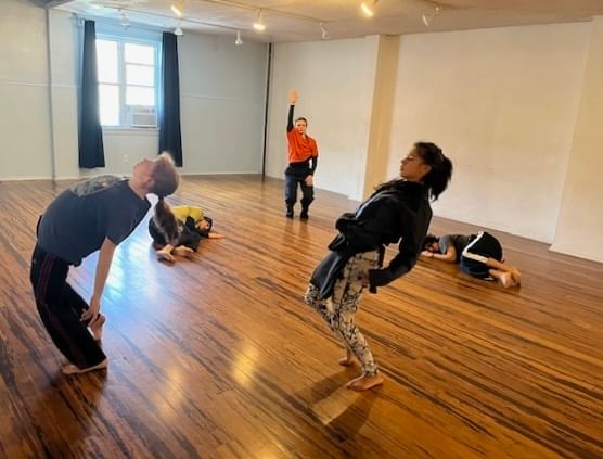 Five performers in relaxed clothing in a brightly lit, open performance space appear to be in a rehearsal