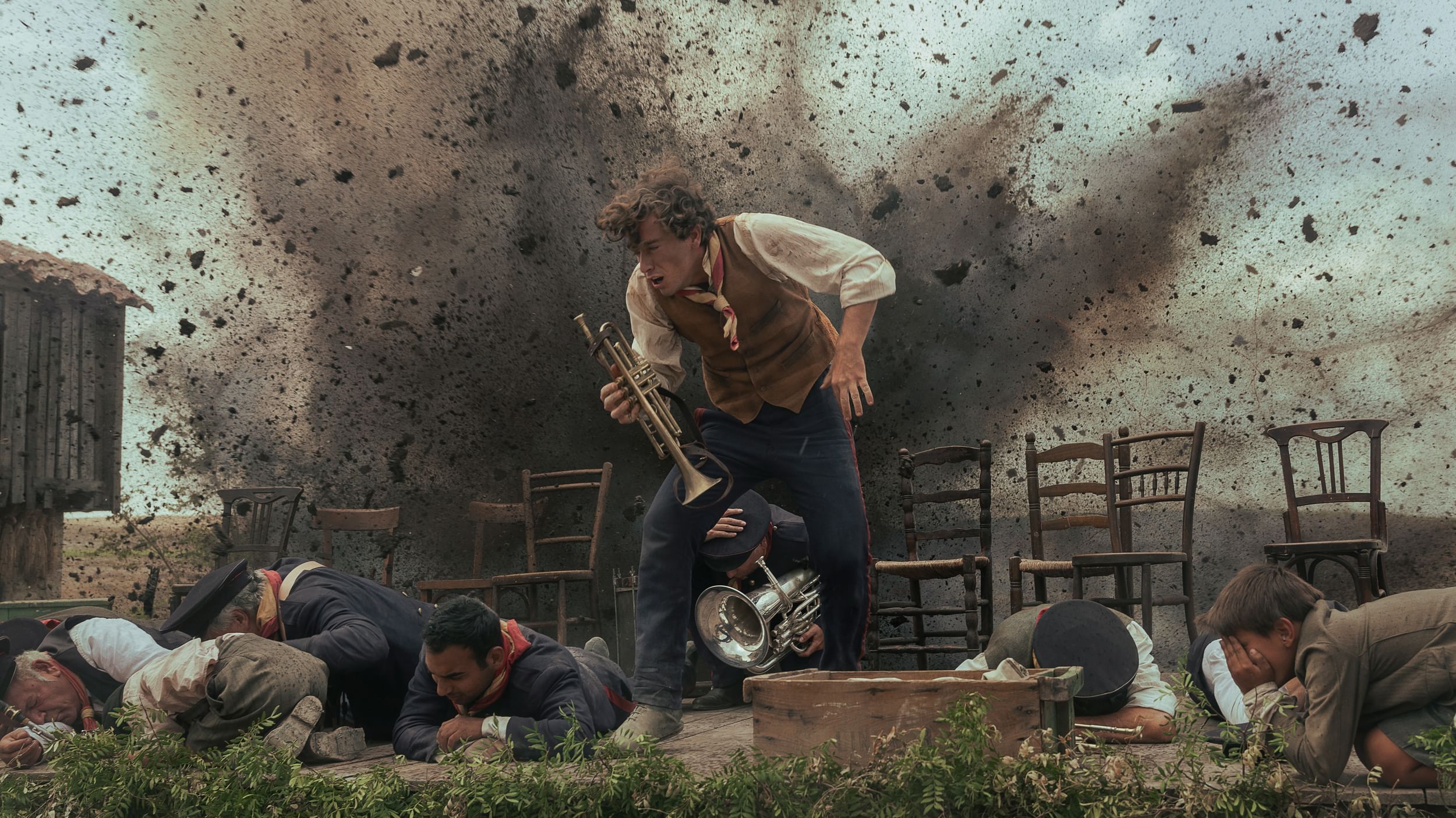 A man holding a trumpet is hit from behind by an explosion as several people in uniform shield themselves on the ground.