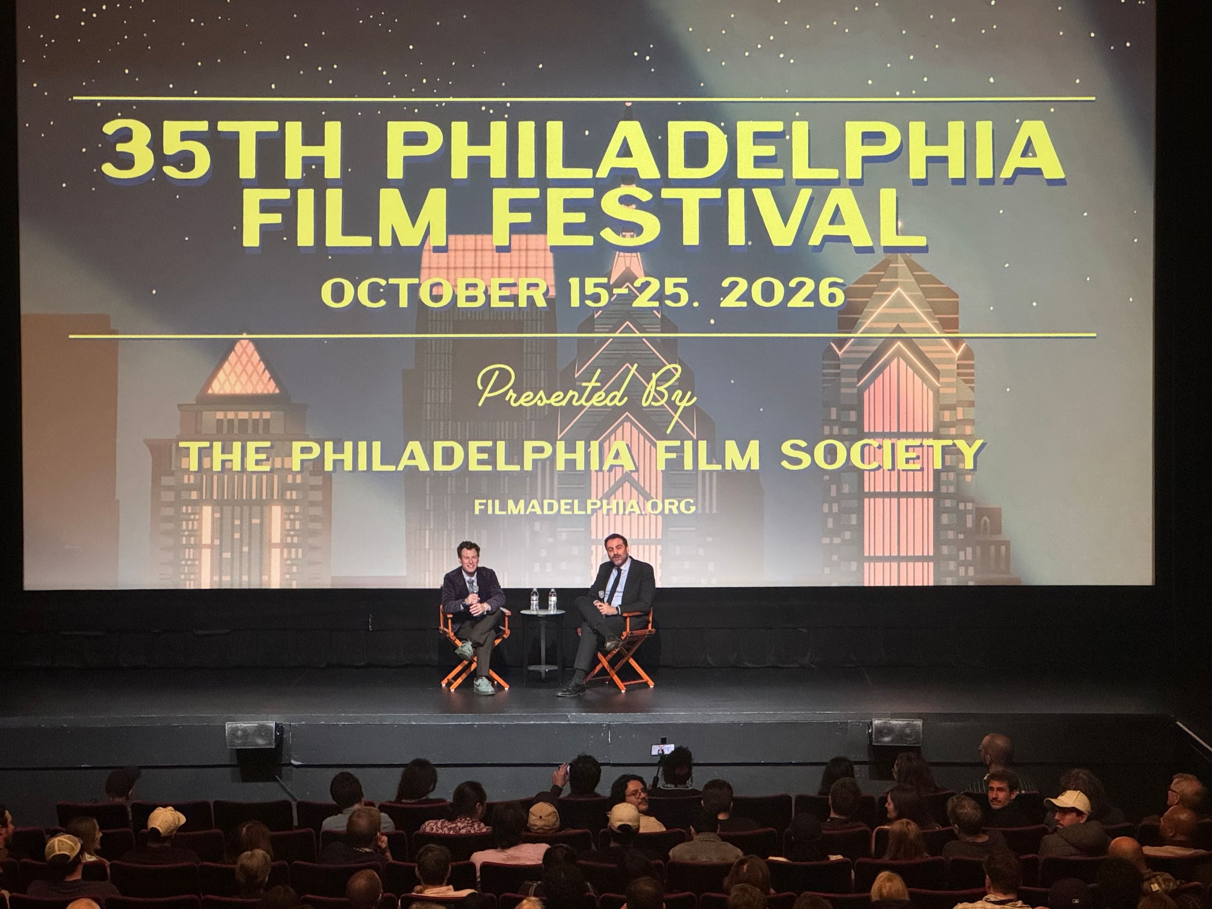 The two men sit opposite each other in director’s chairs onstage, below a large screen with the PFF logo.