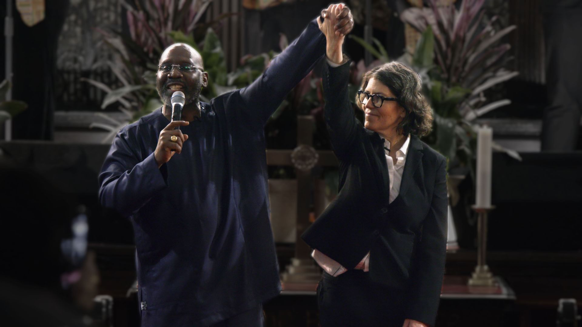 On a chancel, a Black male preacher holding a mic and a white female rabbi join one hand and hold it aloft.