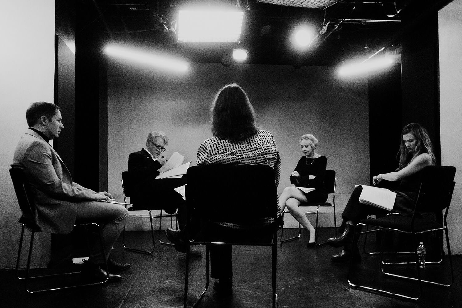 In a stylized, grainy black & white photo, five people sit in chairs in a circle under bright lights, holding scripts.
