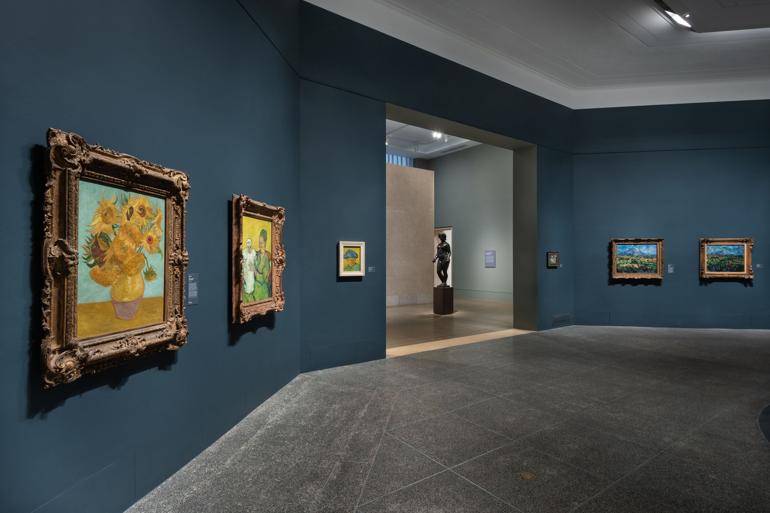 A gallery with angled slate-blue walls displays famous works by Vincent Van Gogh in ornate gold frames.