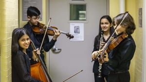 Four grade-schoolers, two boys and two girls, smile in a hallway and hold their instruments: a cello, violins, and clarinet.