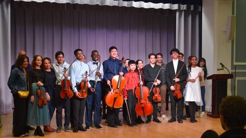 A multi-racial group of about 14 young musicians stands smiling on a small stage, holding instruments, alongside some adults