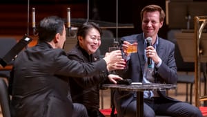 Two performers in all black formal wear cheers with the host also holding a mic at a small table on stage.