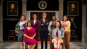 The ensemble of 7 women, in a variety of outfits, pose smiling on the White House set, with the Presidential seal above them.