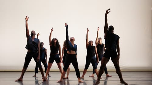 Eight dancers on stage, in mostly all black, all look up with their right arm extended up