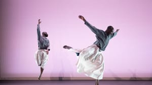 Two Black men in mid-dance, dressed in loose fitted clothes, on a rehearsal stage