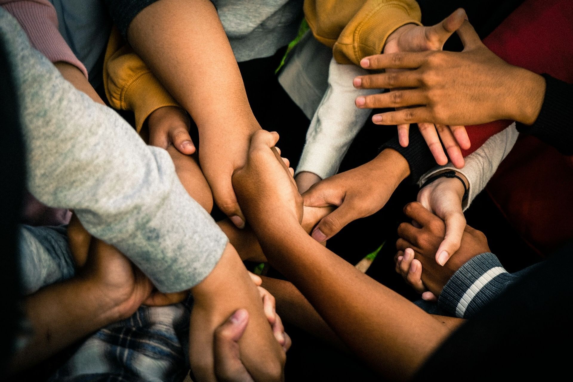 Close-up on many hands of diverse people in a huddle, in different stages of reaching out and grasping each other.