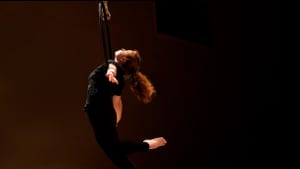 A performer in all black hangs from their shoulders and back a large ring, which hangs in the air