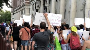 A summer protest city street scene, with a large crowd walking with its back to the camera, holding signs aloft.