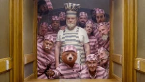A group of 13 older men in matching striped pajamas stand behind Paddington, the CG-animated bear, looking to the camera