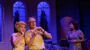 In an opulent house in blue light, Brock and Zack smile together, gesturing with their hands. Quinn is in shadow at right