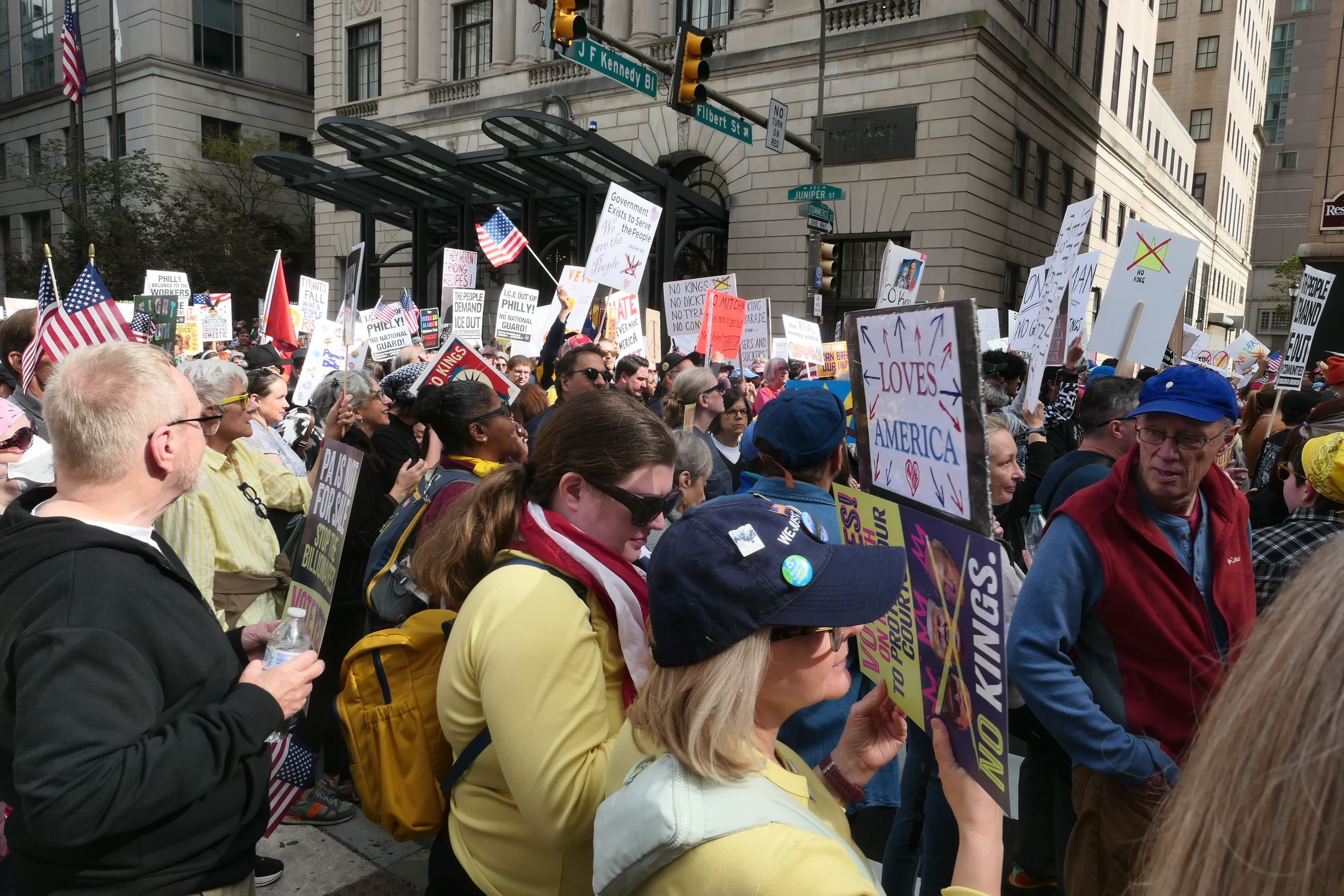 A huge crowd of protestors on a sunny day round the corner of City Hall, holding tons of American flags and handmade signs.
