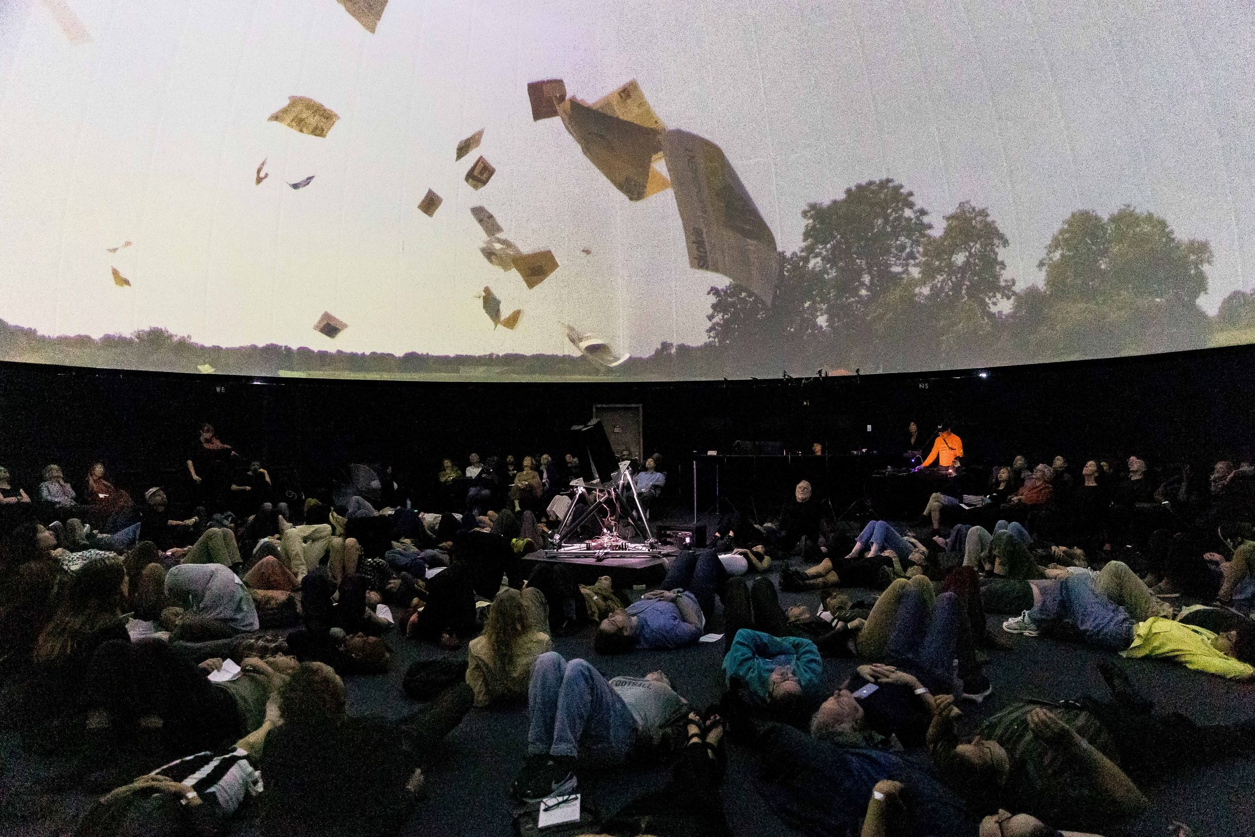 Dozens of people on the floor of the planetarium watch a film of papers whirling in the sky onscreen above them.