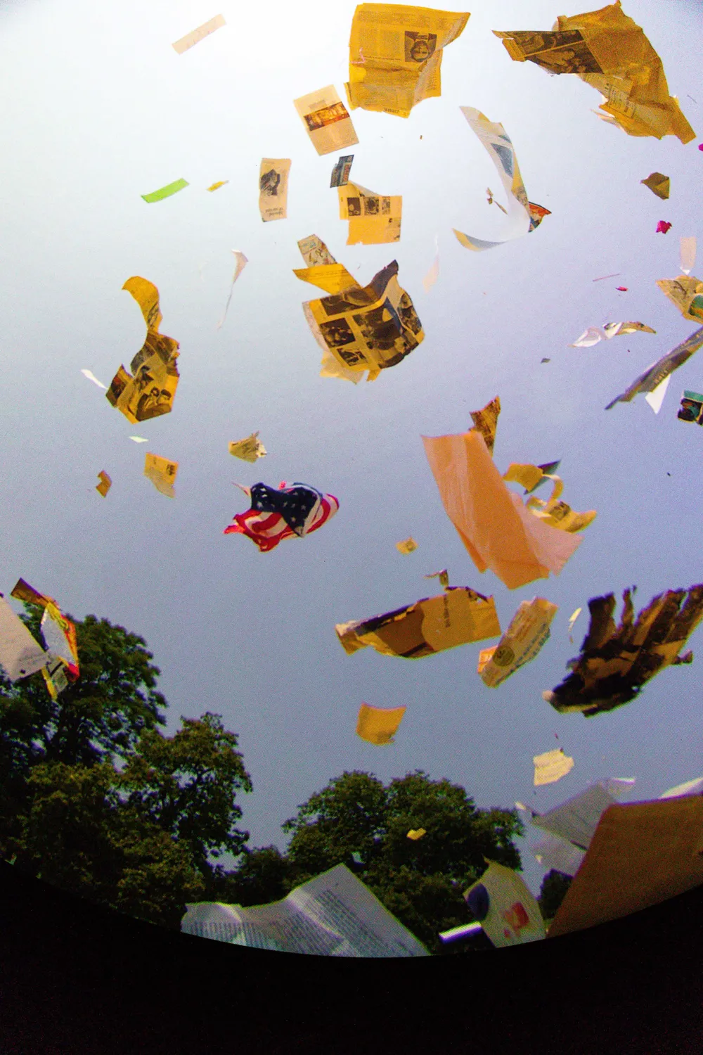 View looking up into a blue sky, above green trees, with a whirl of newspapers and a US flag circling in the air.