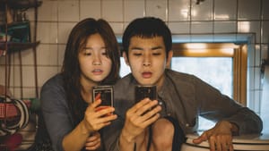 Two Korean people sit close in a bathroom looking at their phones inquisitively