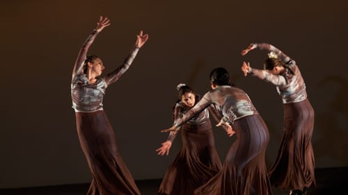 Four dancers in earth-toned outfits with long brown skirts bend gracefully, trailing their arms through the air.