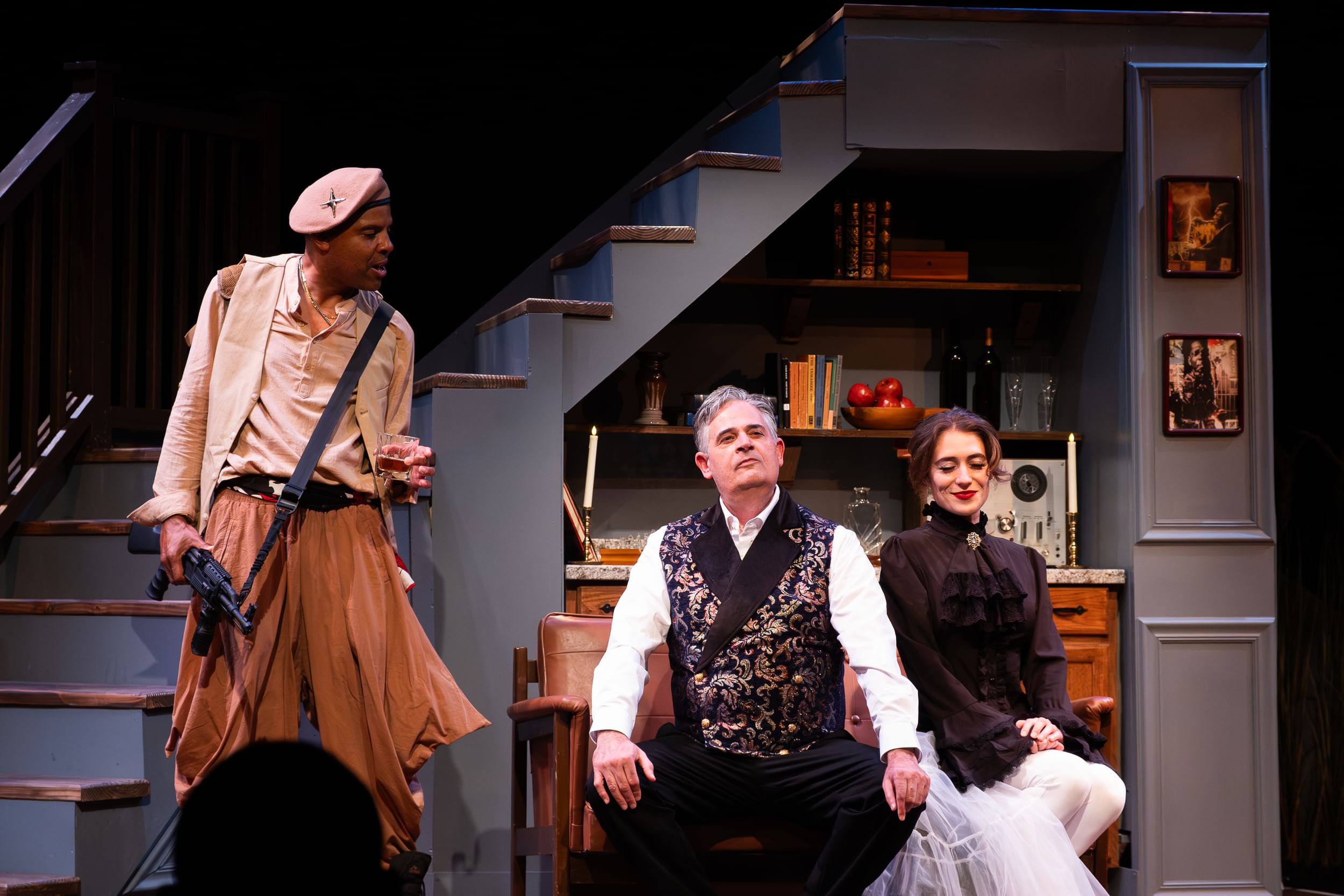 Brown, holding a drink and a rifle and wearing flowing brown clothes, speaks forcefully to Bisgaier & Rose, sitting together.