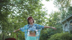 Yelland, in an open plaid button-down, stands smiling with arms spread in a grassy yard. Two breast pumps hang from her chest