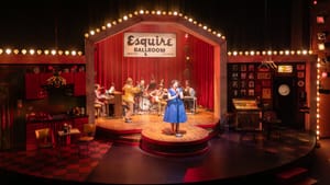 In a blue dress, Lewis-Michelson as Cline sings spotlit on a yellow lightbulb-framed wooden stage in front of the band.