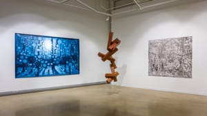 The three artworks: a wooden sculpture hanging in the center and the others on one wall each of the white-walled corner.