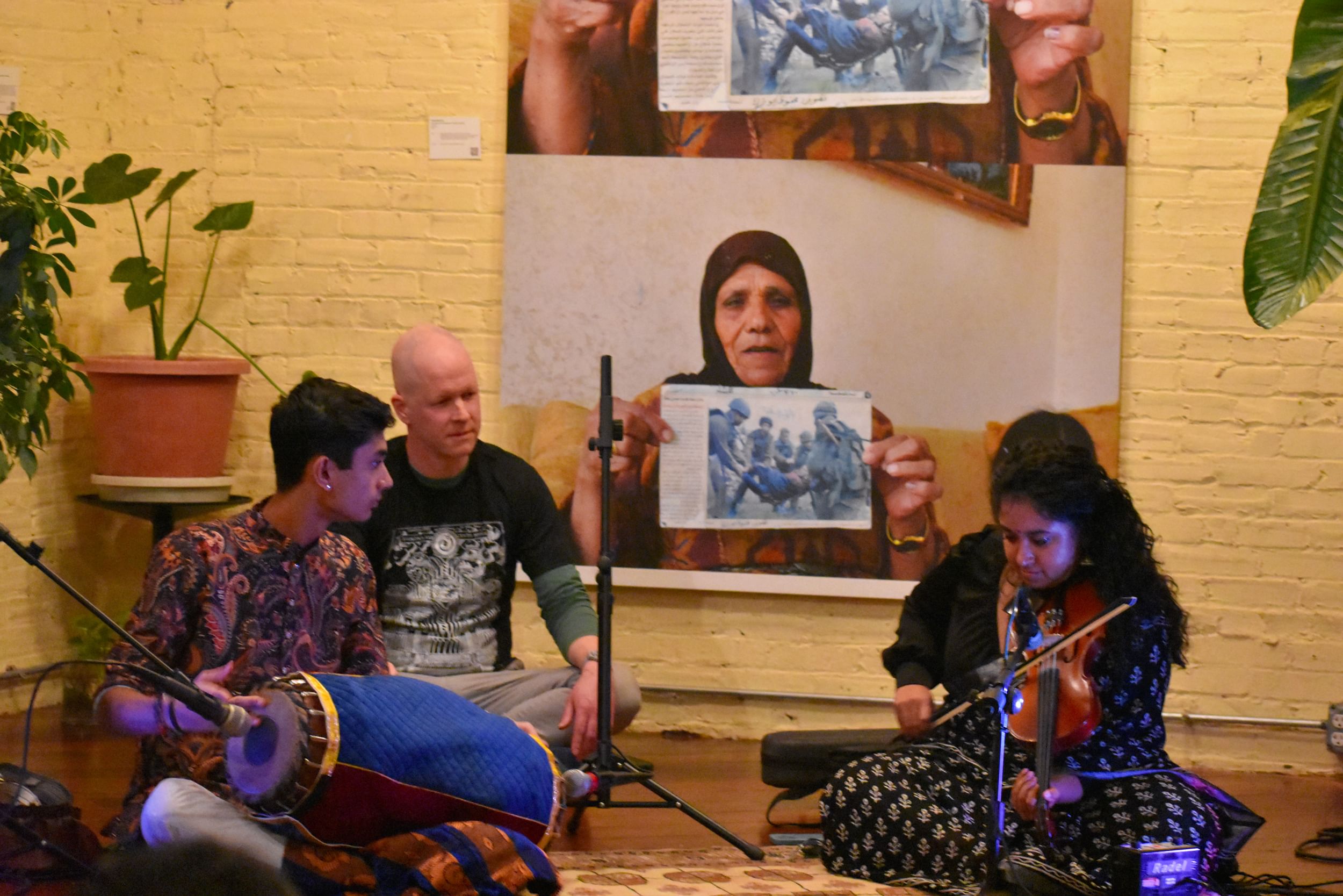 Three people on a low stage, two have instruments, a violin and a drum, in their laps. Third person looks on, observant