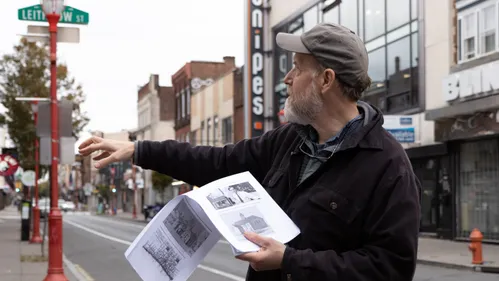 Close-up on Schmitz on South Street, wearing a black jacket and gray baseball hat, gesturing to the left.