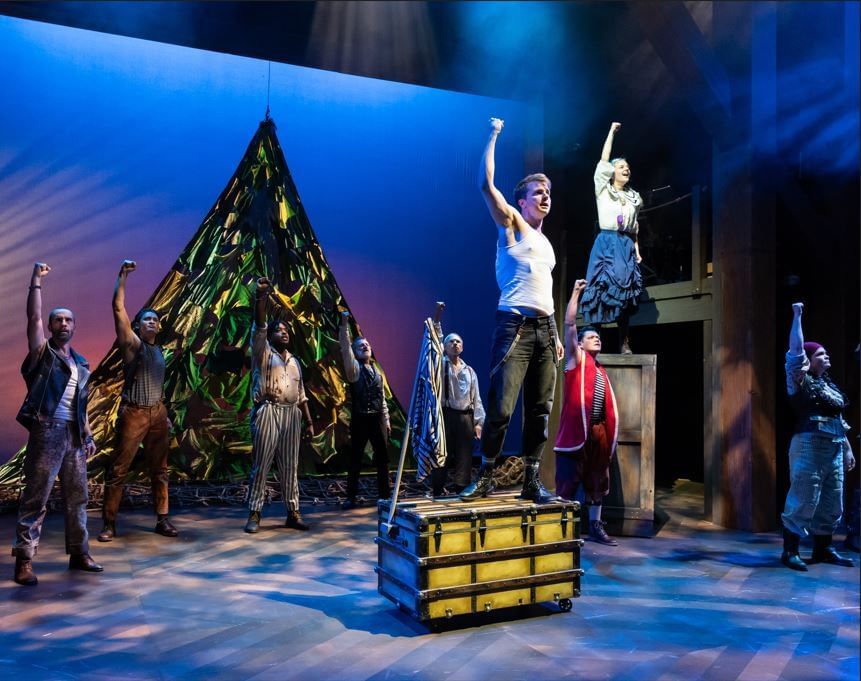 Elmore stands at center stage on a barred wooded chest, with other cast members arrayed around. Everyone raises one fist.