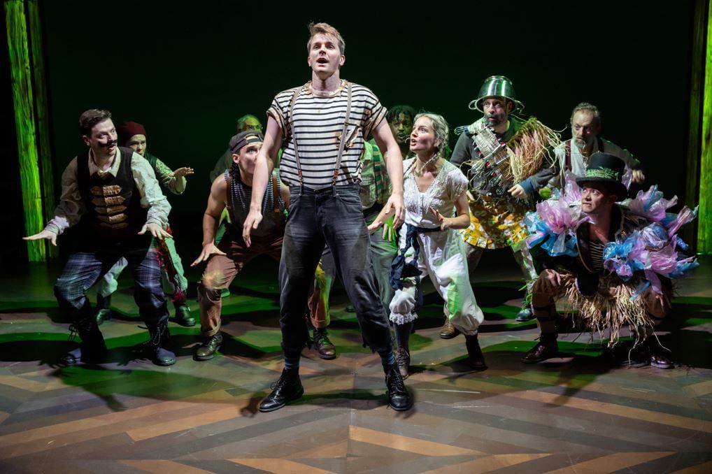 Elmore, in a striped tee & suspenders, sings with a wildly costumed ensemble of about 10 grouped behind him.