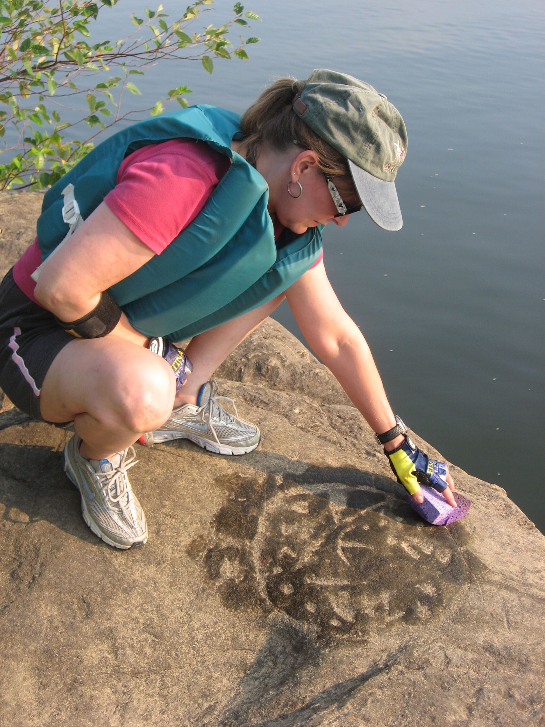 A white woman squats on a brown rock surface, using a wet sponge to reveal an ancient Indigenous carved medicine wheel.