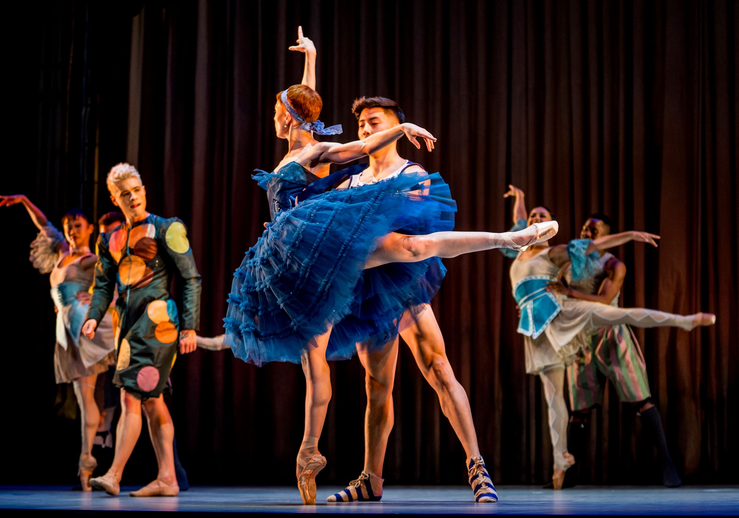 Seven dancers pose in carnival-like costumes, with a ballerina en pointe in a blue taffeta tutu at center, one leg extended