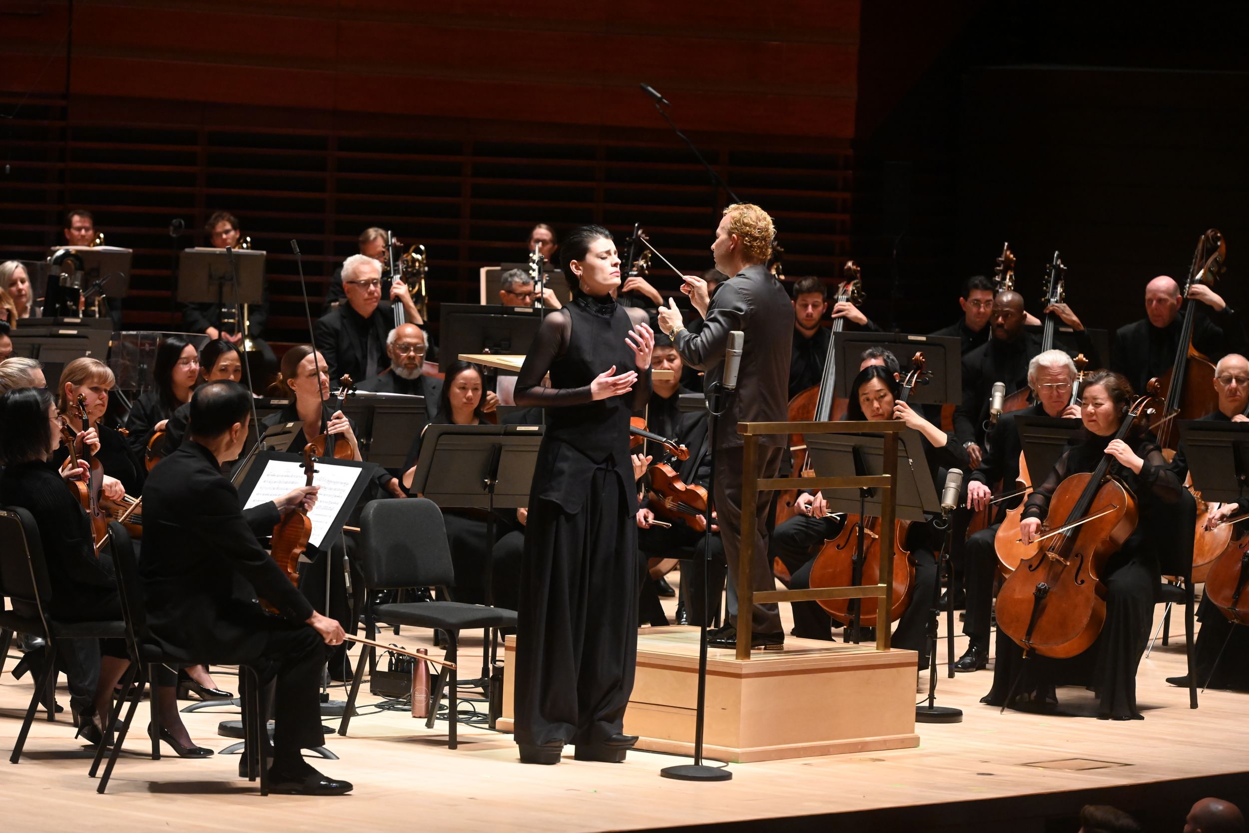D’Angelo, in a black ensemble, sings in front of the orchestra while Yannick, in a gray suit, conducts beside her.