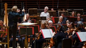 Seen from behind Nézet-Séguin energetically conducts DiDonato, with short silver hair & white dress, standing among musicians