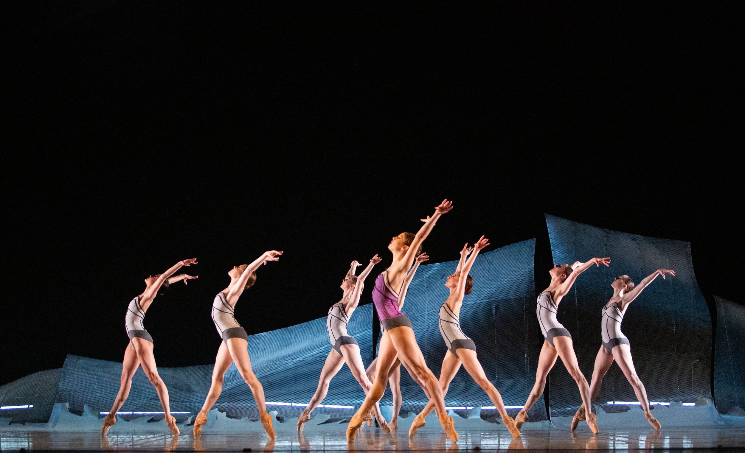 Nine female dancers en pointe, in structured leotards, pose in profile with legs apart like open scissors, arms flying behind