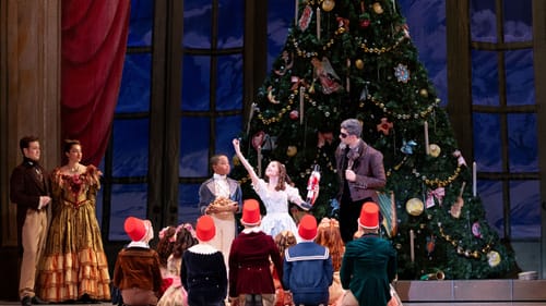 Young dancers sit around a girl in a frilly dress, gleefully holding a nutcracker doll, all in front of a huge Christmas tree